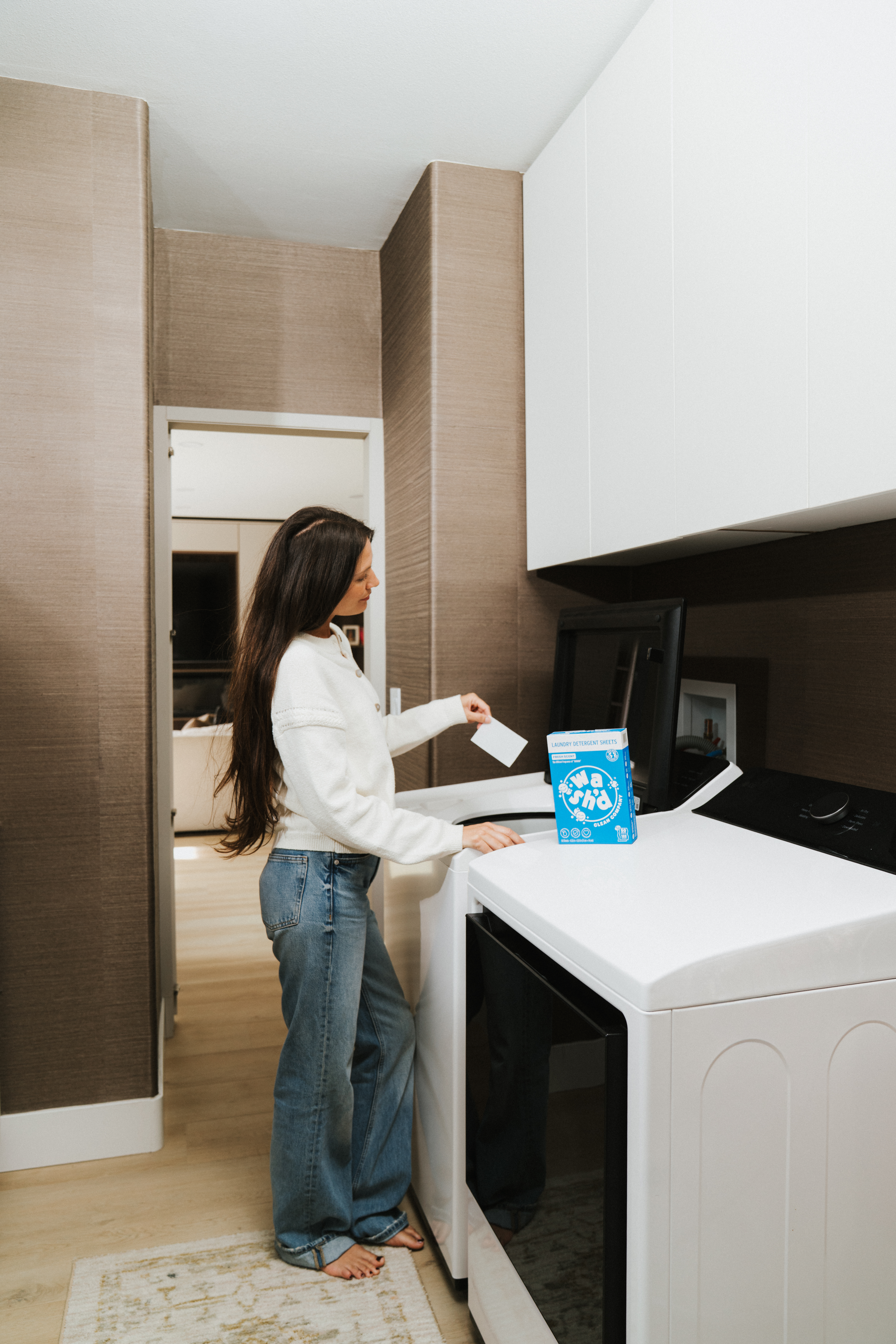 A woman in a white sweater stands at a washing machine in a modern kitchen