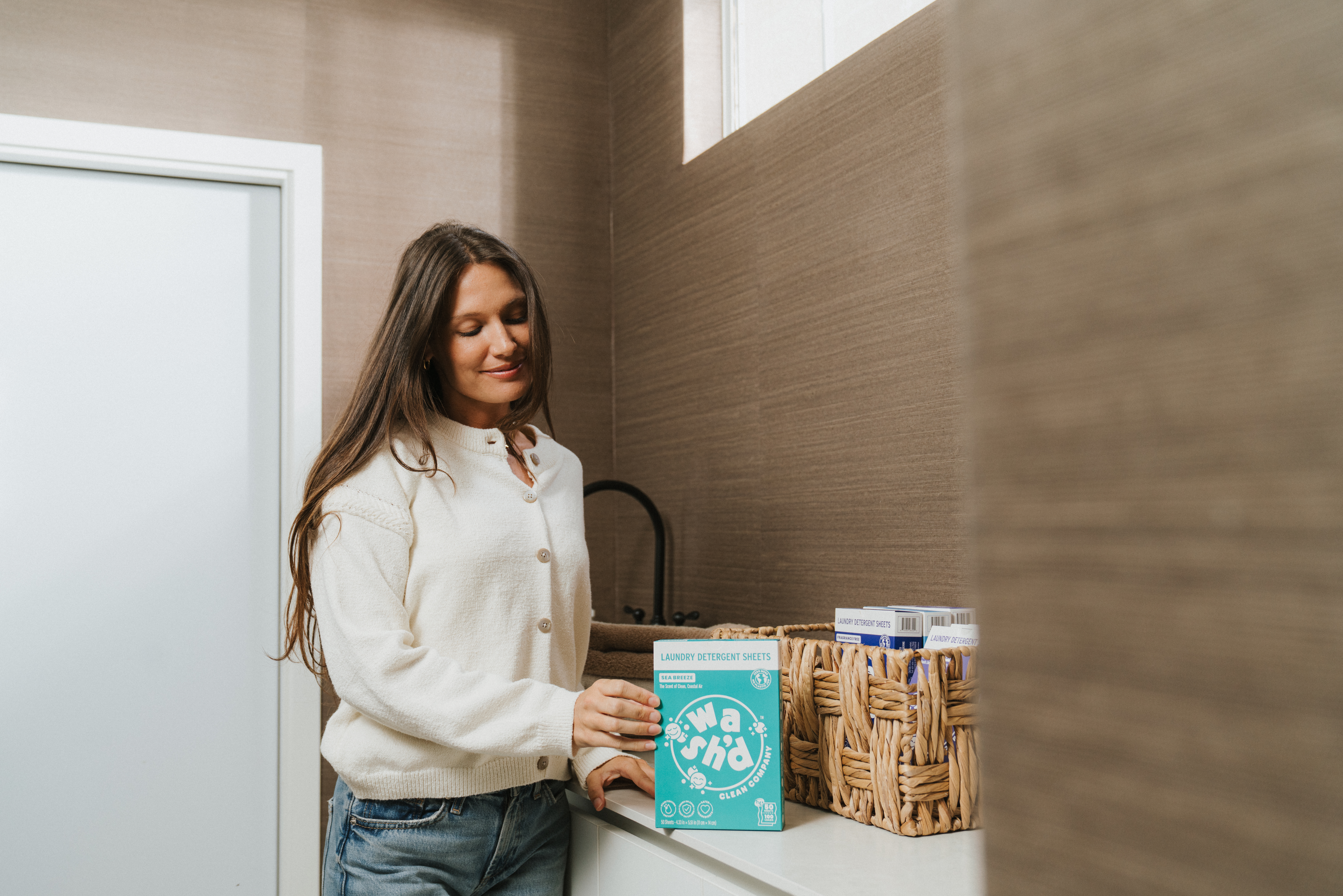 A woman in a white sweater holds a Wash'd detergent sheets box beside a woven basket of products on a white counter