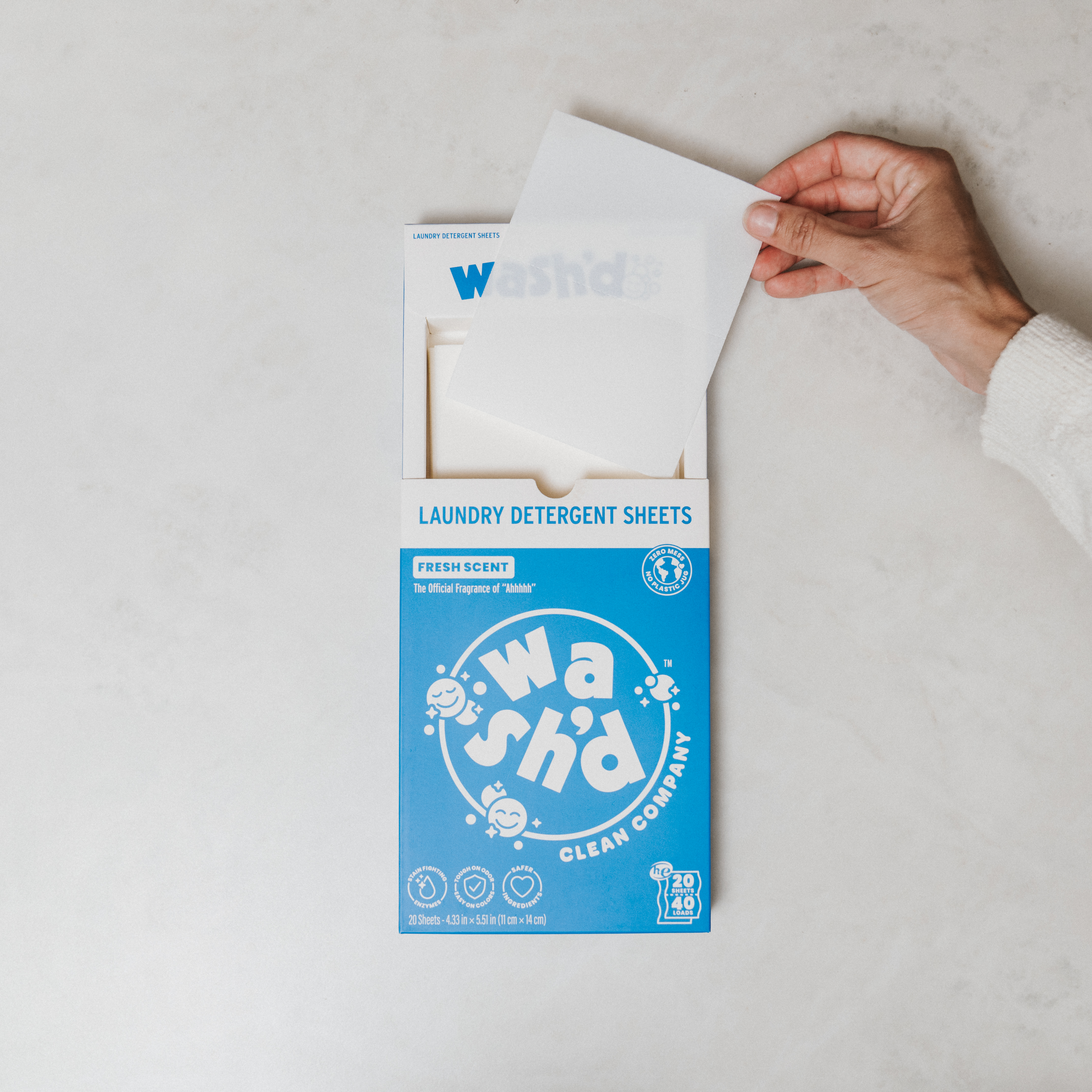 A hand pulls a white laundry detergent sheet from an open Wash'd Fresh Scent box