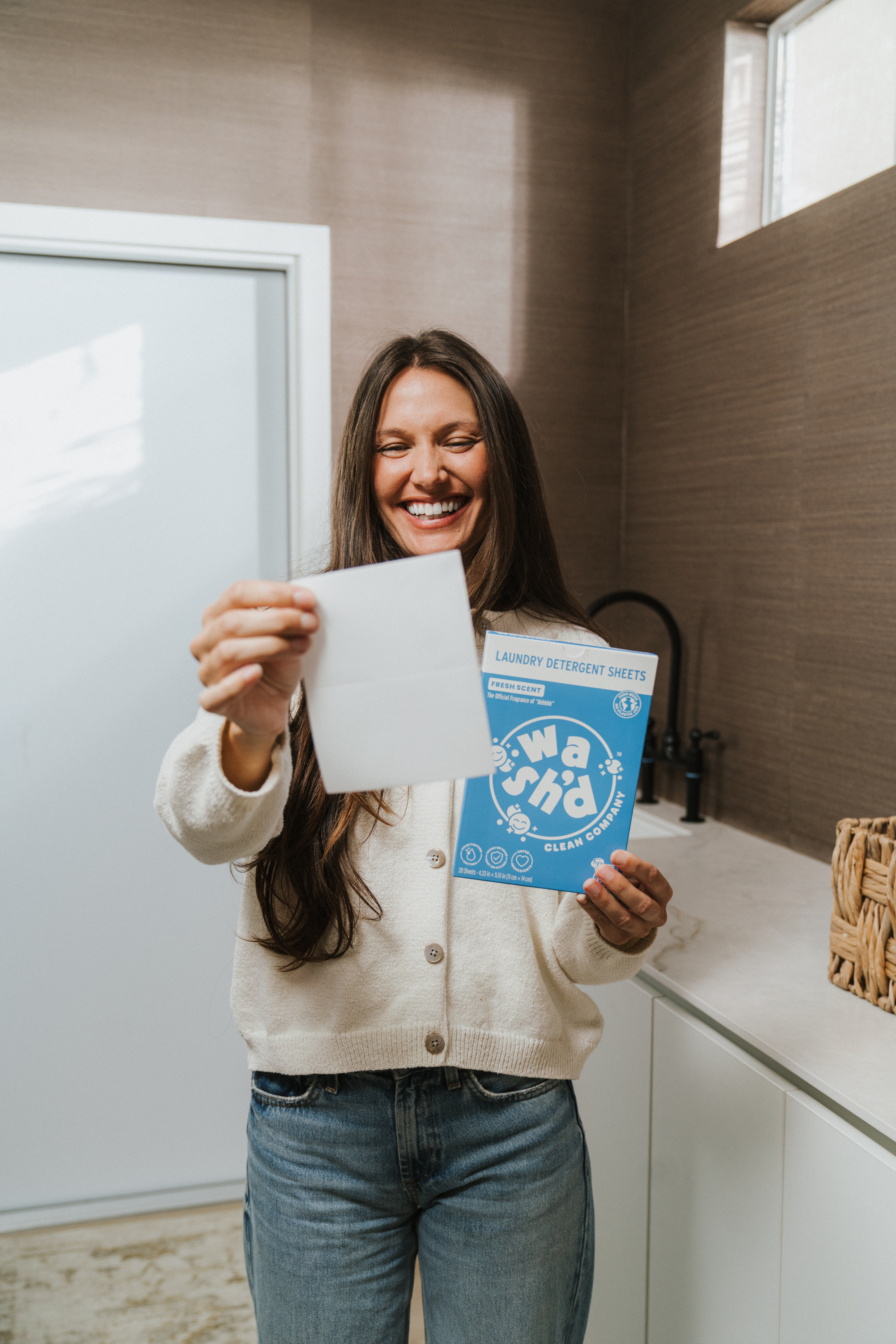 Woman holding a single Wash'd laundry detergent sheet beside the blue Wash'd packaging in a bright modern bathroom